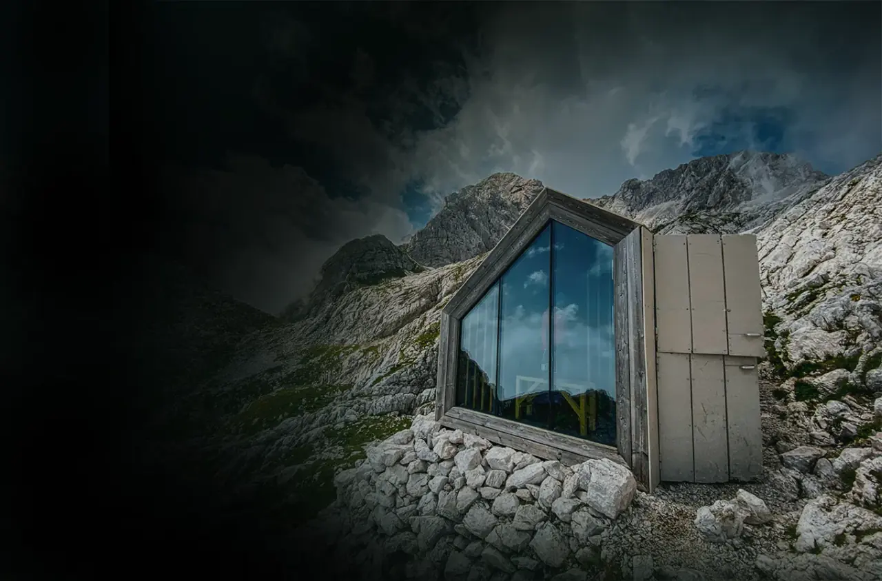 A glass-fronted alpine bivouac on a drystone base among limestone scree under a storm sky, ridges of the Kamnik Alps rising behind it.